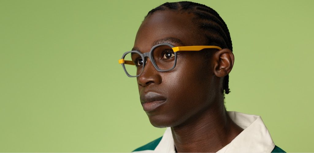 Close-up side-profile portrait of a man wearing Scout Mateo bold geometric eyeglasses with a grey front and bright yellow temples against a light green background. His hair is styled in neat braids pulled back from his face and he's wearing a collared top