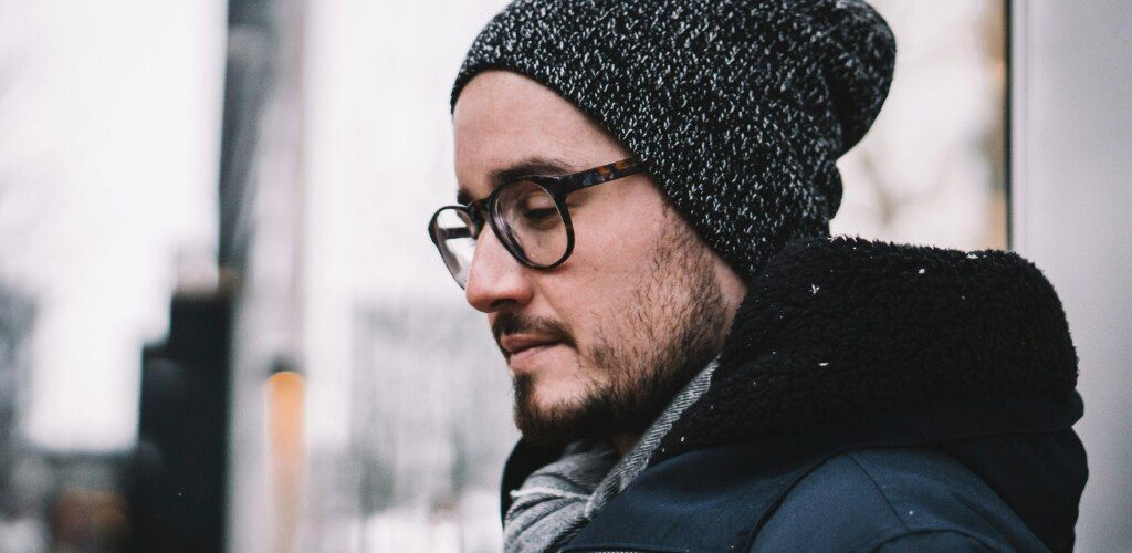 A man with stubble wearing round tortoiseshell glasses, a grey beanie and a winter coat outdoors in the snow.