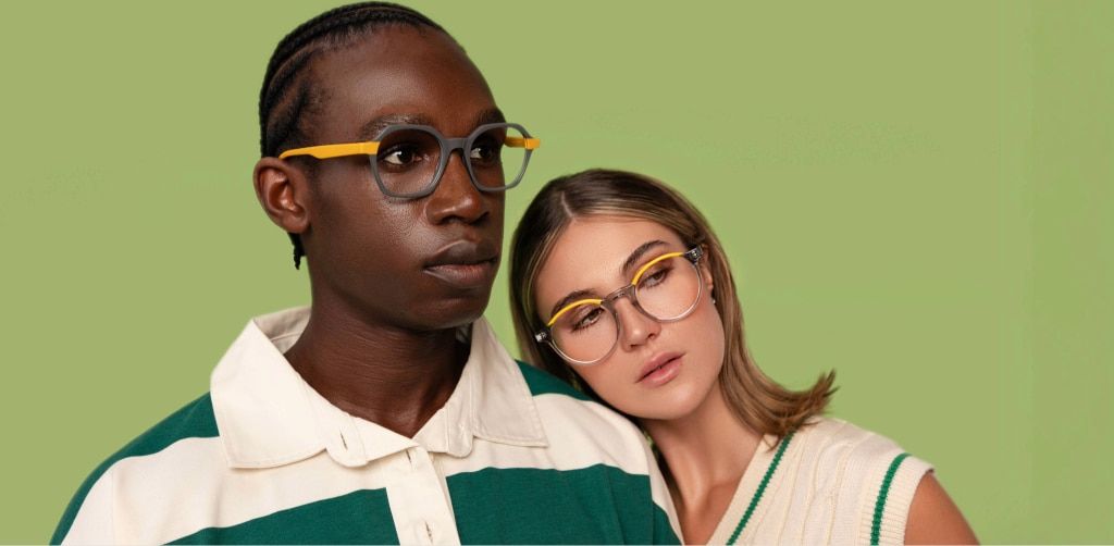 Two models wearing coordinated round glasses with yellow temple and brow details, posing against a soft green background, styled in neutral and green tones with a relaxed, contemporary look.