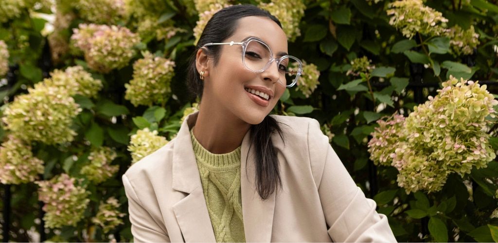 Woman with dark hair in a ponytail wearing transparent oversized glasses, smiling outdoors in front of green foliage, styled in neutral tones with a blazer and knit top.