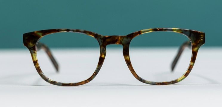 Front-facing product shot of The Collection Andi tortoiseshell glasses resting on a white surface against a teal background. The frame features a classic rectangular shape with slightly rounded edges and a distinctive keyhole bridge. The mottled brown, am