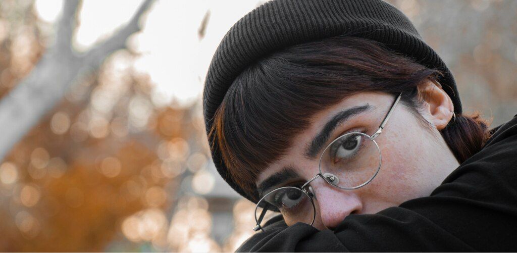 Person wearing a black beanie, round wire-frame glasses and a black top, peeking over their arm against a blurred autumn backdrop — 90s-chic eyewear inspiration.