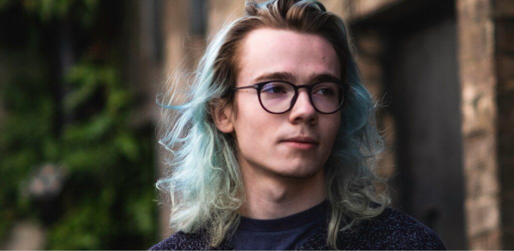 Man with long blue-highlighted blonde hair wearing round black glasses and a dark navy sweater, standing against a blurred urban backdrop — grunge-revival eyewear inspiration.