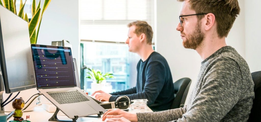 Two men sitting in a bright office, looking at dual computer screens