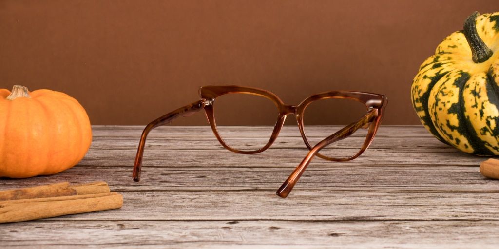 A pair of acetate brown gradient cat-eye glasses are resting open, facing away on a wooden surface. Surrounding them are pumpkins and cinnamon sticks.