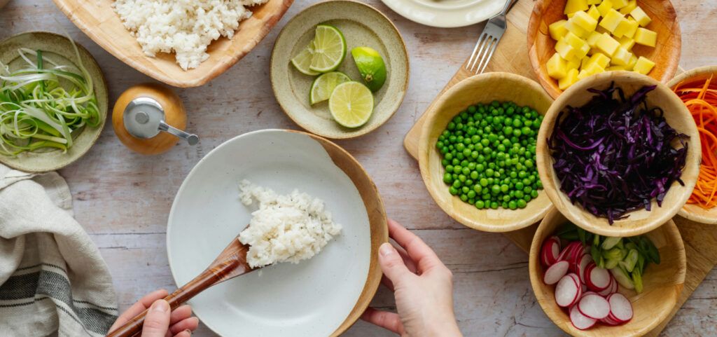 A high-angled shot of a selection of wooden and porcelain bowls and plates, containing healthy, brightly coloured foods and vegetables, including carrots, sliced cabbage, peas, radish slices, potato cubes, lime slices, spring onion and white rice. At the