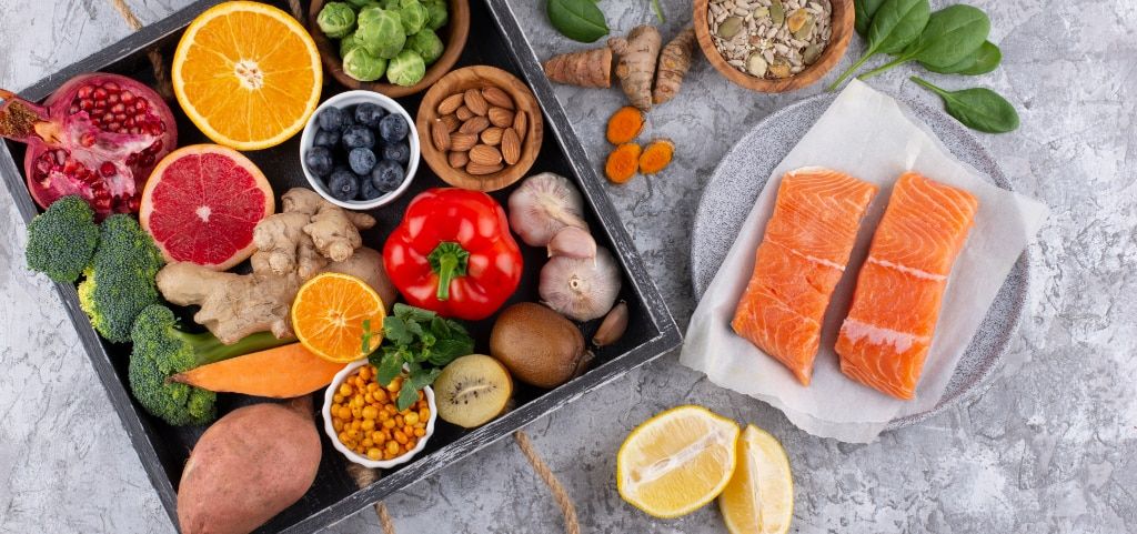 A selection of healthy foods on a grey marble surface, including two salmon fillets on grey paper and a grey plate, alongside a dark-grey wooden tray with rope handles filled with oranges, blueberries, almonds, Brussels sprouts, and other produce.