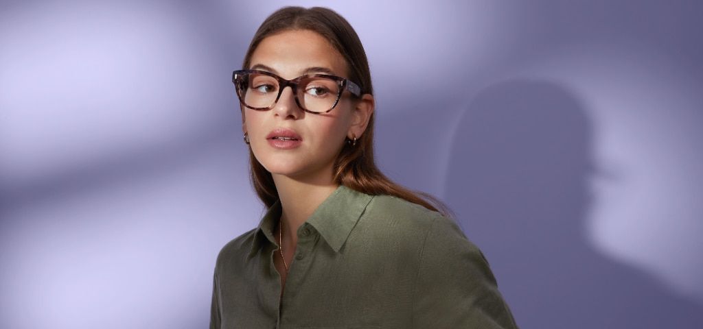 Woman with long brown hair wearing tortoiseshell prescription glasses and an olive-green shirt looking to the side against a soft purple background.