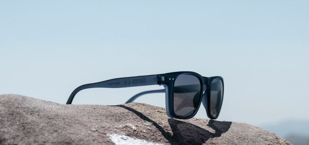 A pair of dark acetate sunglasses perched open on a grey rock with the blue sky overhead.