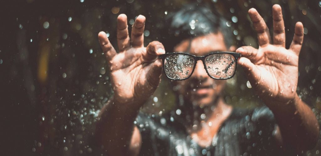 A person holding a pair of black rectangular eyeglasses in front of them in rainy conditions. Water droplets are visible on the lenses, partially obscuring the view through them. Their thumbs and forefingers grip the frame while the other fingers are spre