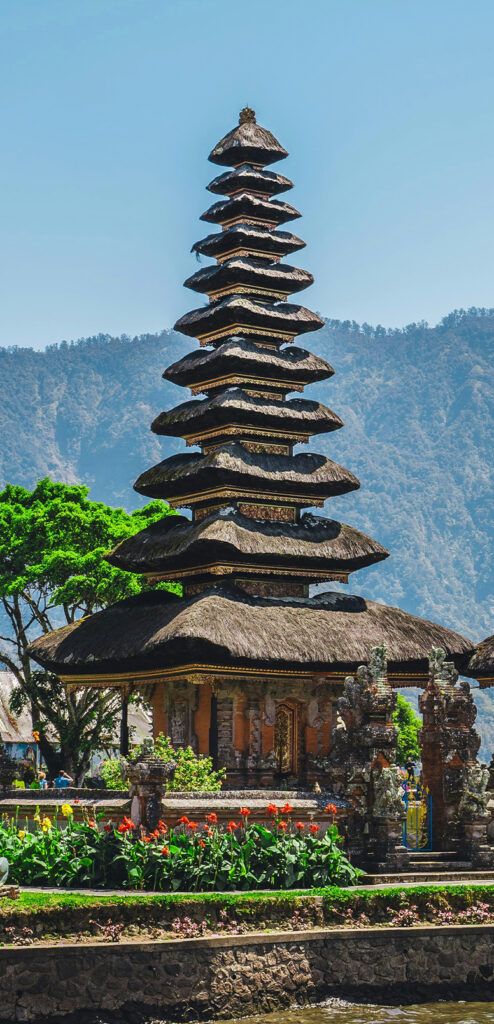 A photo of a temple in Bali. It has a multi-tiered, thatched-roof and is located on the waterfront. There are mountains visible among mist in the background.
