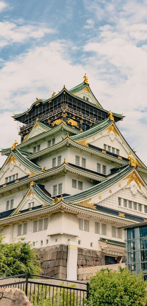 A photo of Osaka Castle's main tower, a five-story building with emerald-coloured roofs, ornate detailing and gold trim. Green foliage surrounds the site.
