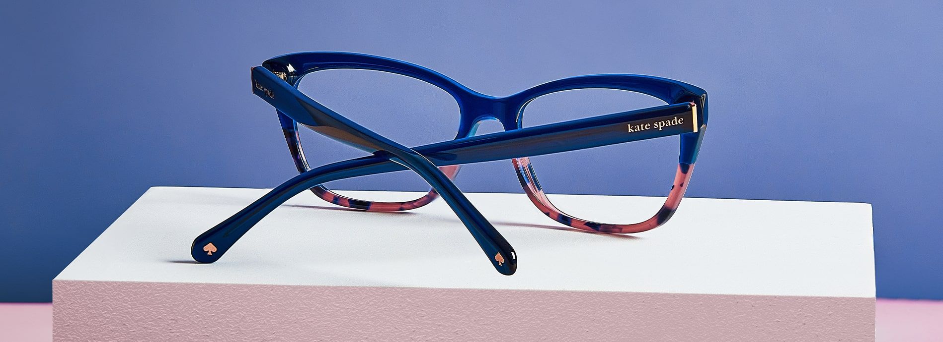 Close-up of blue to brown tortoiseshell ombre style Kate Spade frames from the reverse showing the Kate Spade logo and spade emblem on the arms, positioned on a white block with light blue background