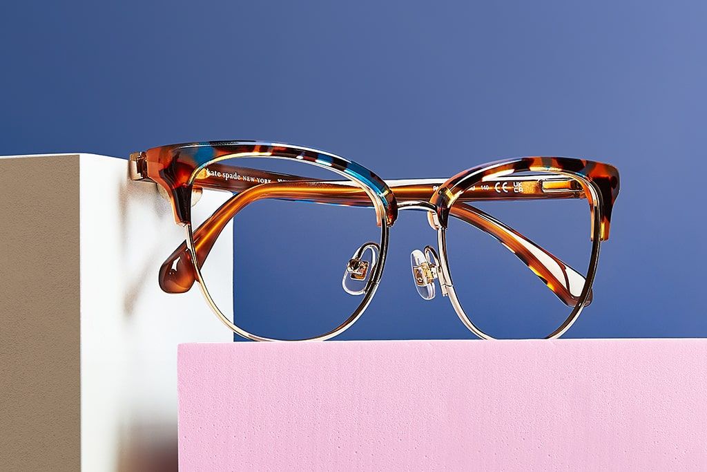 Close-up of clubmaster style brown tortoiseshell and gold metal Kate Spade frames facing the right, positioned on a pink and white block with light blue background
