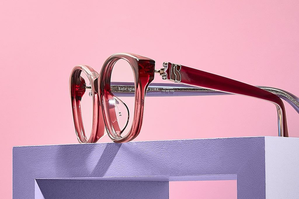 Close-up of red acetate Kate Spade frames showing the details on the frame arm facing left, positioned on a purple block with pink background