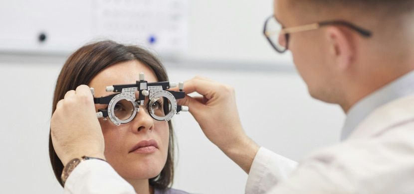 Optician conducting an eye exam on a woman with medium brown hair, using trial lenses to check vision and update prescription glasses.
