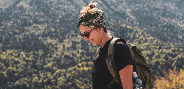 A young lady with her hair tied up with a black and white bandana around her head. She appears to be hiking in a bright outdoor environment. The lady is smiling brightly, wearing black sunglasses, a t-shirt and a green backpack with a water bottle visible