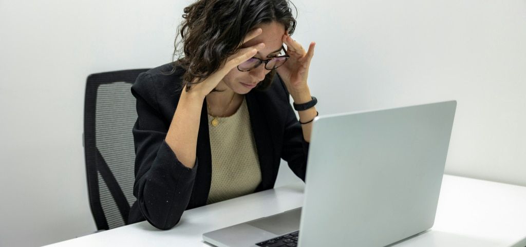 A lady sitting in a bright office space, sat at a desk looking at a laptop and holding her head in her hands
