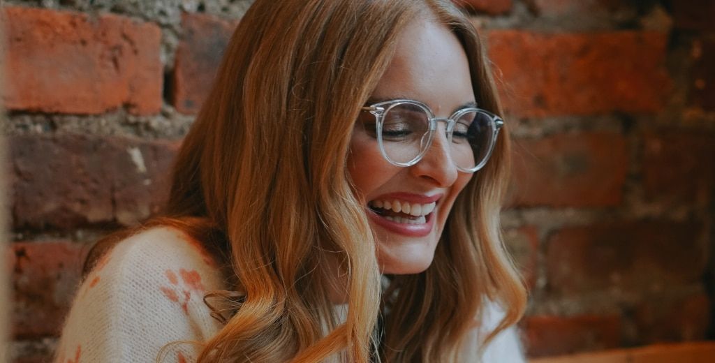 A blonde-haired woman laughing in a bright space with a brick wall as the backdrop. She appears to be looking down at something. She is wearing a pair of transparent glasses and a white cotton jumper with flowers on it.