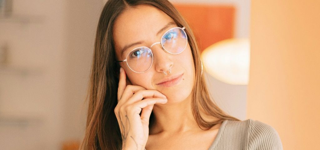 A close-up of a young lady with a nose piercing, wearing round metal glasses and a grey top. She has long light-brown hair and is resting her face on her right hand. She appears to be in a bright indoor setting.