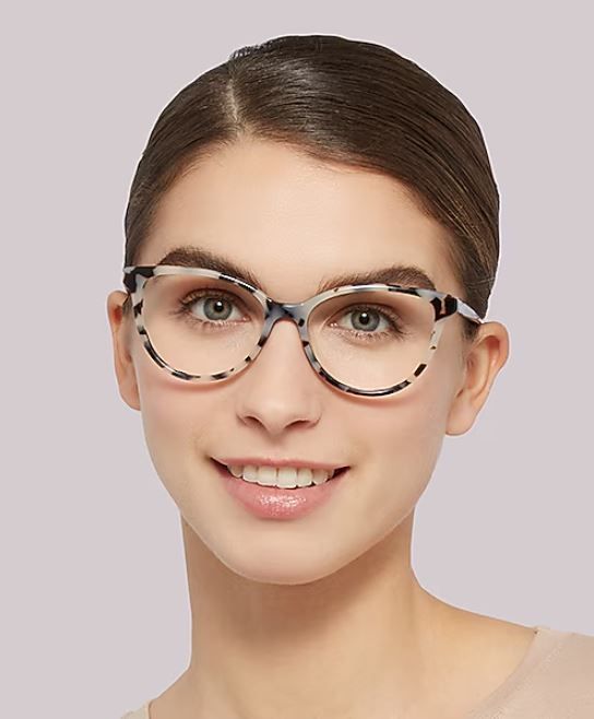 Front-facing studio portrait of a young woman wearing a beige top and cat-eye tortoiseshell-style eyeglasses, with brown hair neatly pulled back and a soft neutral background.