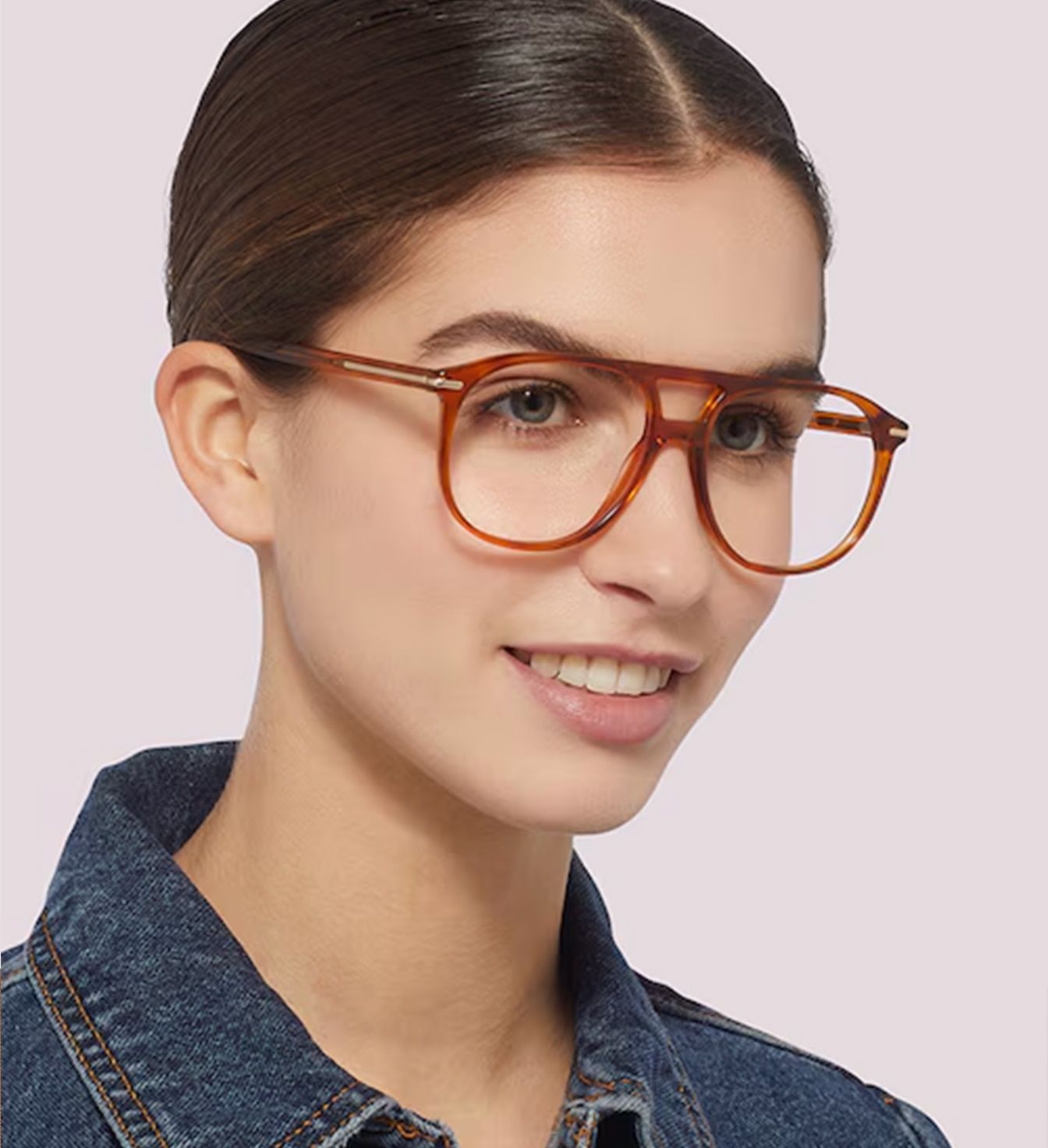 A young lady in a pair of large honey-coloured pilot glasses. Her face is turned to her left side. She has brown hair which is tied up and slicked back. The lady appears to be wearing a denim shirt which is just visible.