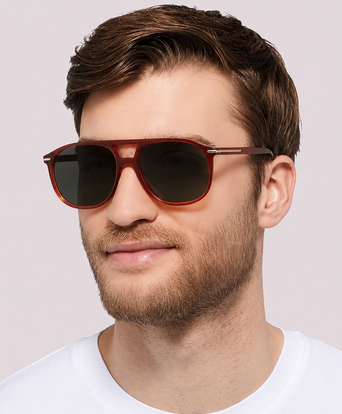 A man with short brown hair styled in a tousled side quiff and light stubble wears large orange aviator-style sunglasses and a white t-shirt, looking slightly to the side with a neutral expression against a plain background.