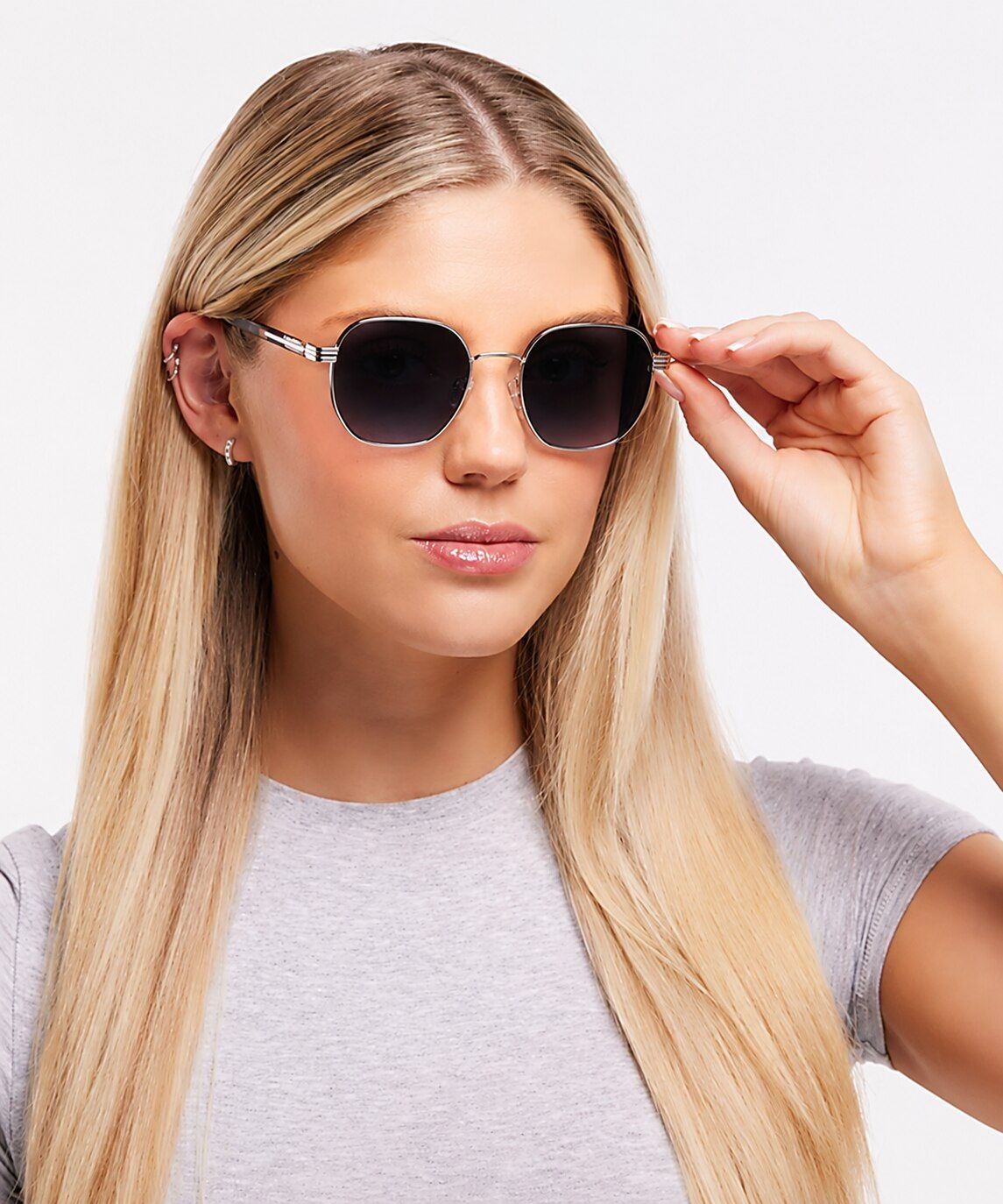 A woman with long blonde hair wears oversized geometric sunglasses with gold-toned frames. She gently touches the temple of the sunglasses with one hand while gazing slightly off-camera. The portrait is set against a plain light background, creating a cle