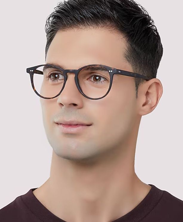 A man with dark, styled hair wears a brown V-neck top and round tortoiseshell eyeglasses while looking slightly upward and to the side. The clean portrait, set against a soft neutral background, highlights the lightweight round frames and gives a smart, c