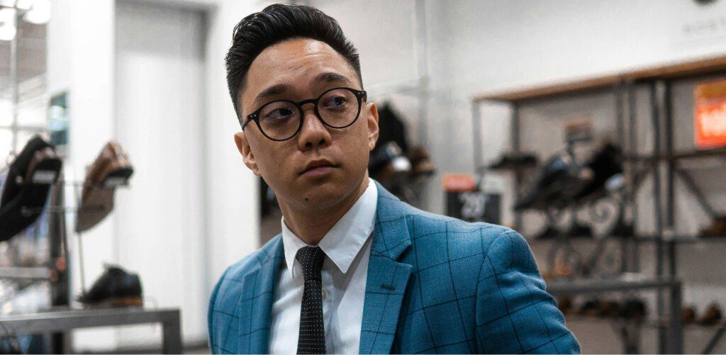Man with dark hair wearing round black glasses and a blue checked suit jacket in a retail store, showcasing a comfortable and secure low-bridge fit.