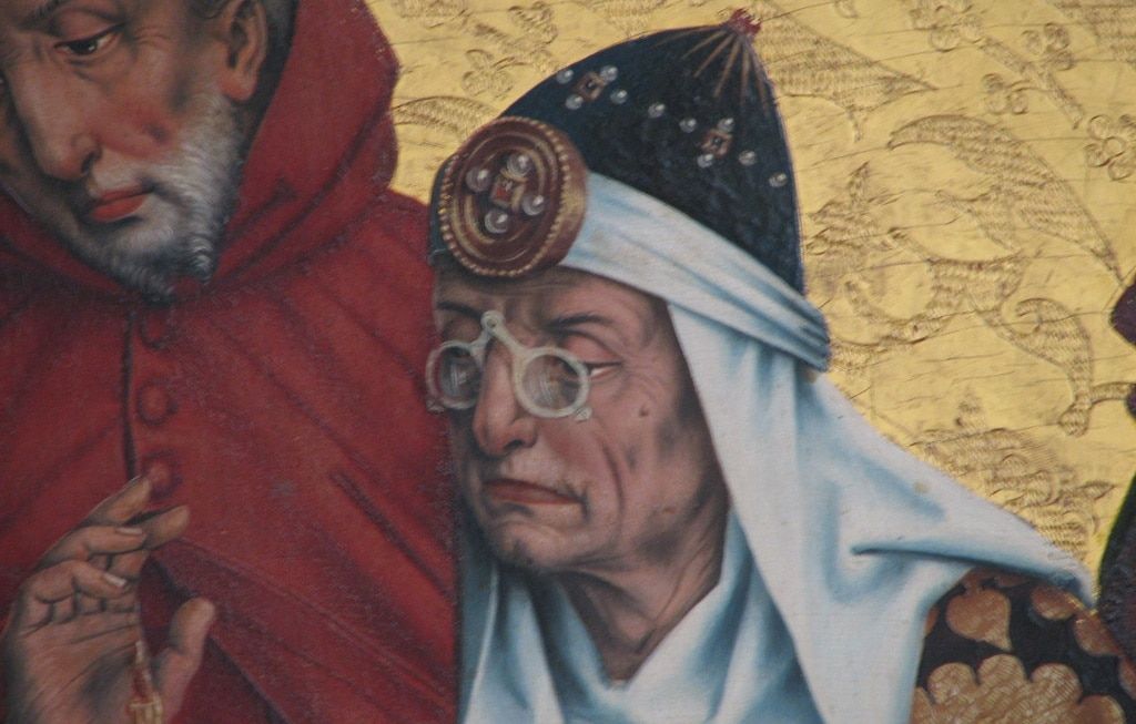 A Medieval illustration showing an old man with a Middle Eastern head-dress wearing scissor glasses on his nose.