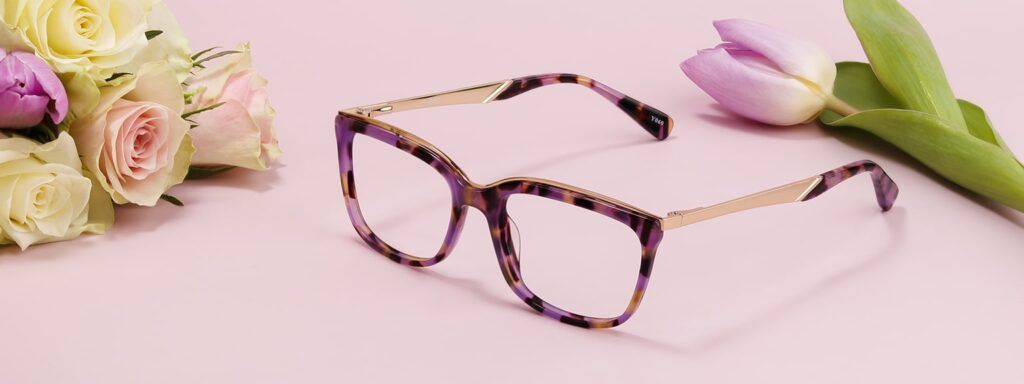 Purple Havana rectangular women’s glasses with gold metal temples displayed on a pink background with a bunch of roses and a tulip, styled for a Mother’s Day gift guide.