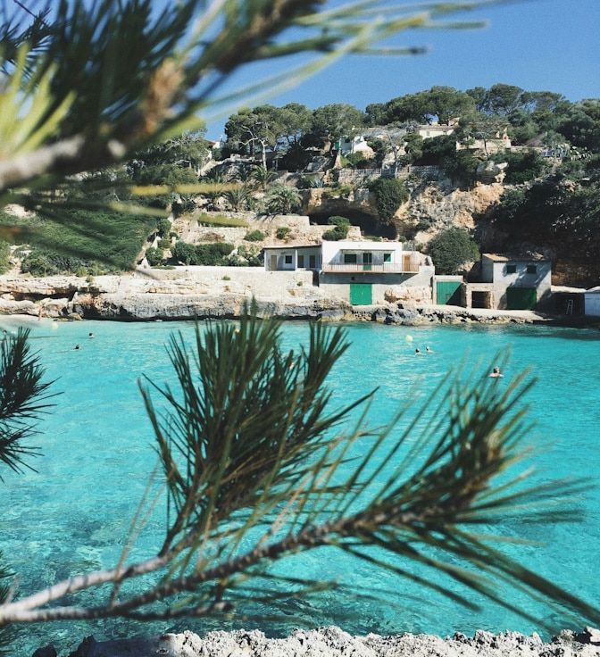 A rocky beach in Mallorca with trees and small buildings close to the bright turquoise seaside
