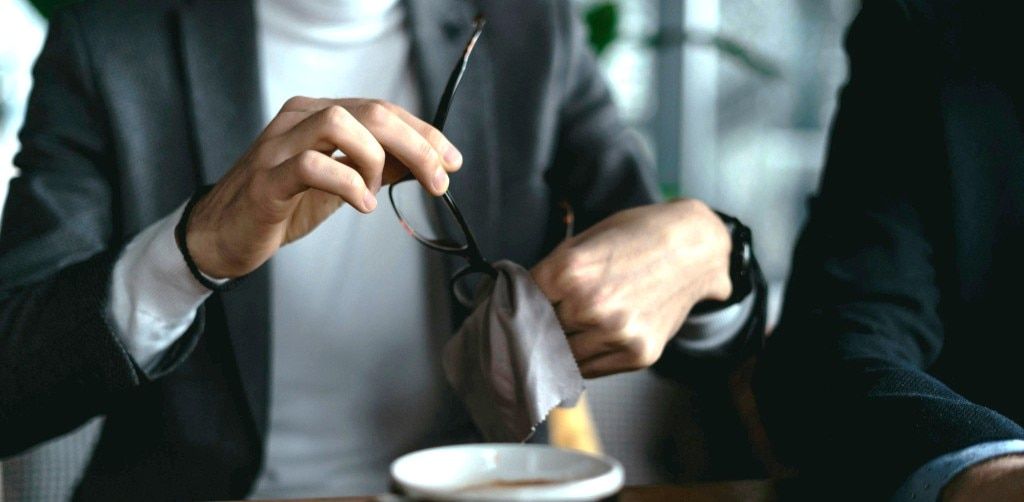 Close up of a man's hands cleaning a pair of tortoiseshell glasses with a microfibre cloth while seated at a table with a coffee cup.