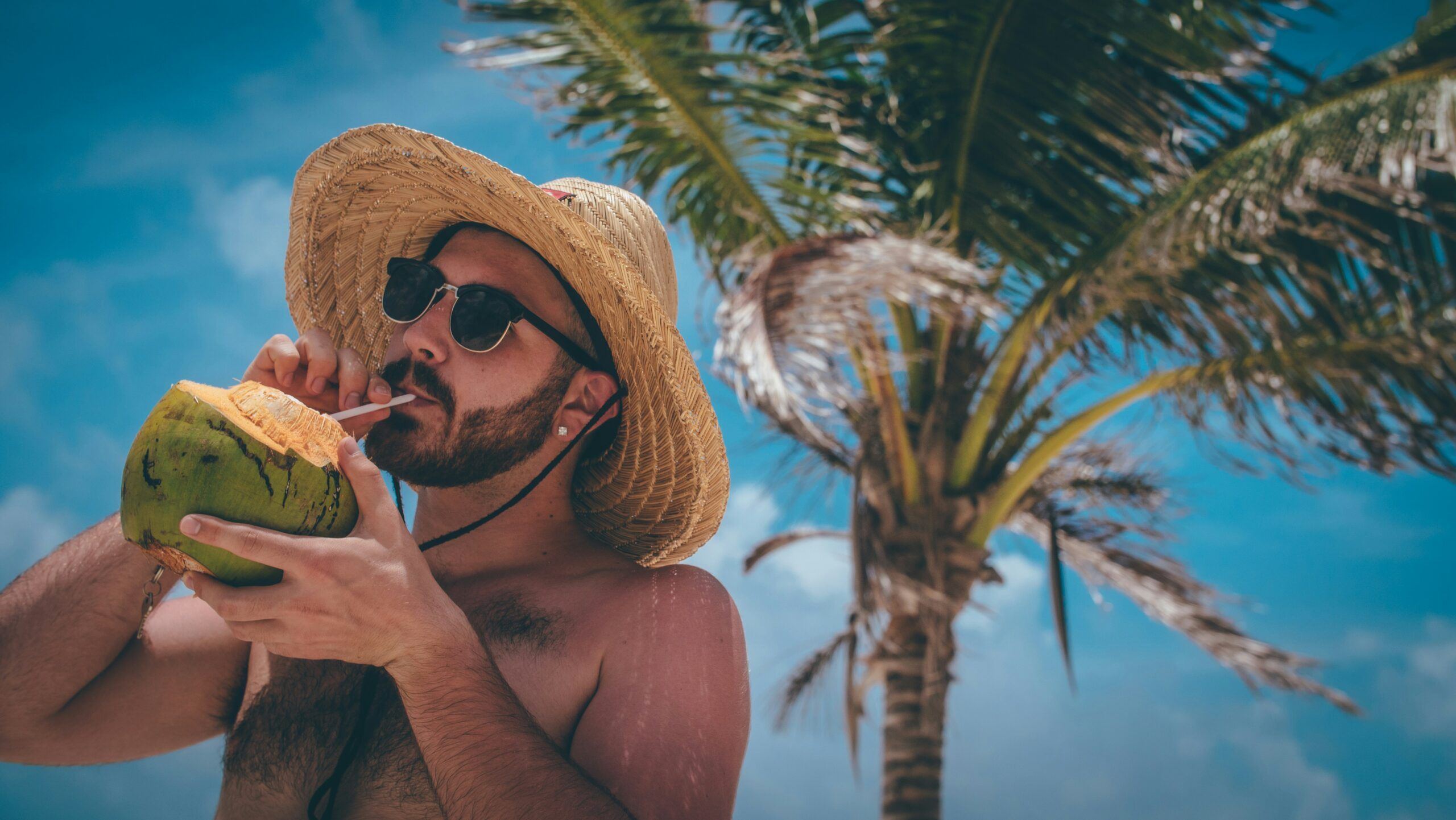 A young man with dark hair and a neatly trimmed beard stands in a tropical setting. He is topless, wearing a wide-brimmed straw sun hat and dark sunglasses with gold metal rims. In his hand, he holds a coconut, sipping from it through a straw.