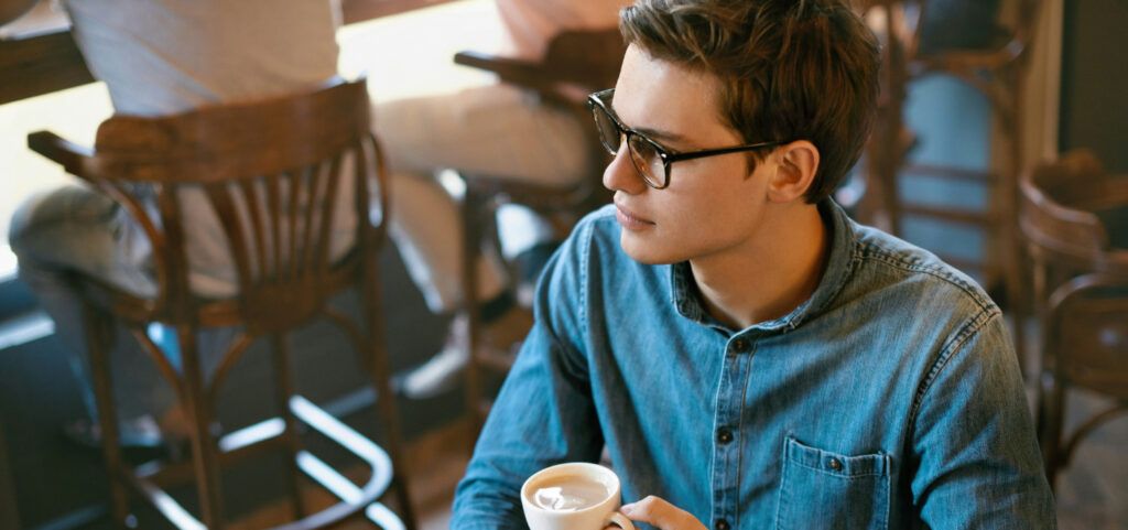 A close shot of a young man, sat comfortably in a coffee shop with people sitting on stools in the background. He has short brown hair and is wearing a pair of black square glasses and a denim shirt. He is holding a coffee cup in one hand and looking away