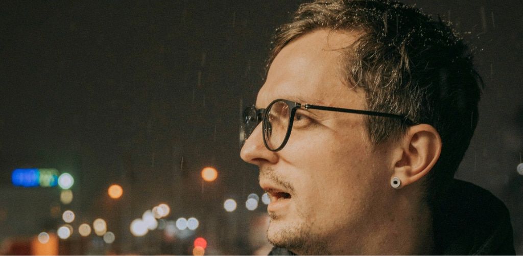 A man with short grey hair wearing round black glasses stands outdoors at night in the rain. Raindrops are visible falling around him, and city lights blur into colourful bokeh in the background. Despite the wet conditions, his lenses appear relatively cl