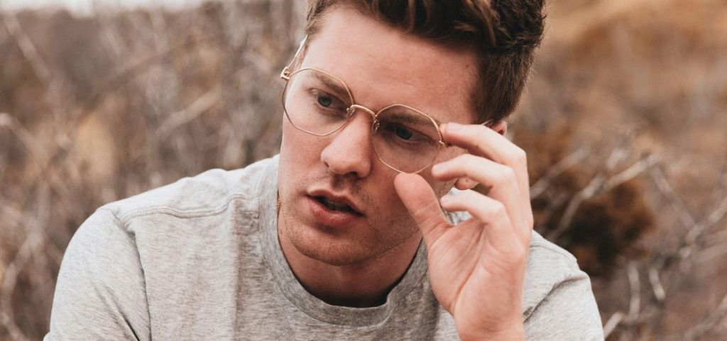 A close up of a young man with auburn hair. He is wearing gold-tone metal geometric glasses and a grey tee. His left hand is lightly clasping the frames of his glasses as he looks off into the distance. He appears to be sitting in a field with dry grass j