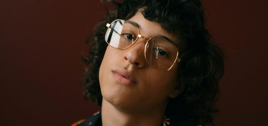 A close-up of a young man with curly 70-style dark hair. His head is tilted dramatically to one side and he is in a dark setting. He is wearing a pair of square glasses made of gold metal.