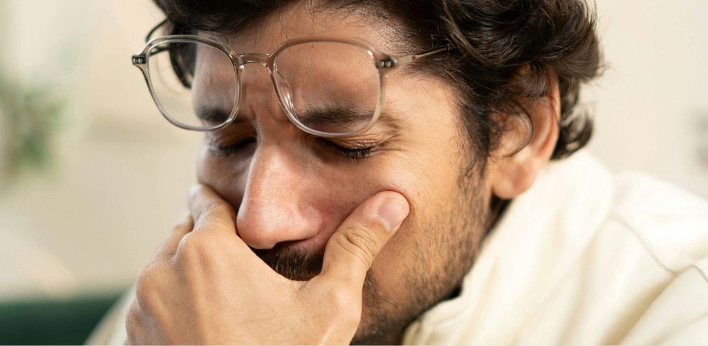 Man with short dark curly hair, wearing a white fleece and clear glasses on his head with his eyes closed, showing eye strain and fatigue, holding face in discomfort.
