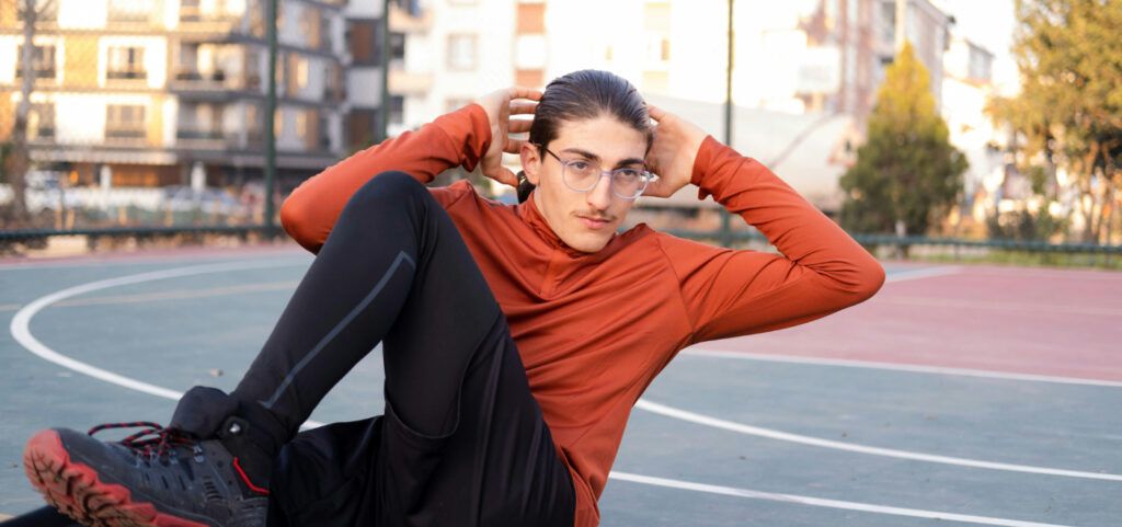 A young man stretching on an outdoor running track. He appears to be performing press-ups with two hands held behind his head and his left knee bent in the air. He has his hair tied back in a ponytail, is wearing clear glasses, a red sweater, black jogger