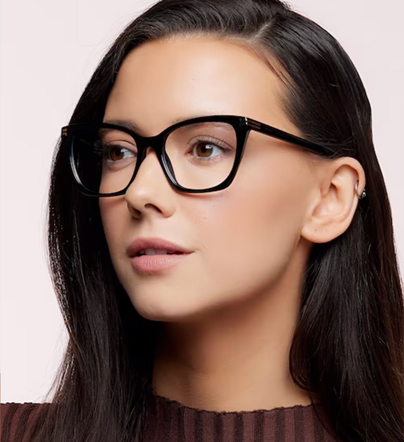 A young lady with medium-length dark hair is wearing a pair of black cat-eye glasses. Her face is turned to her right side. She is also wearing a brown jumper which is just visible.