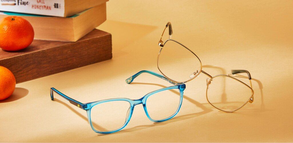 Two pairs of eyeglasses are styled on a warm-toned yellow surface. One pair has a bold, translucent blue square frame with New Balance branding and a more modern, eye-catching look, while the other features thin, gold metal cat-eye-shaped rims for a light