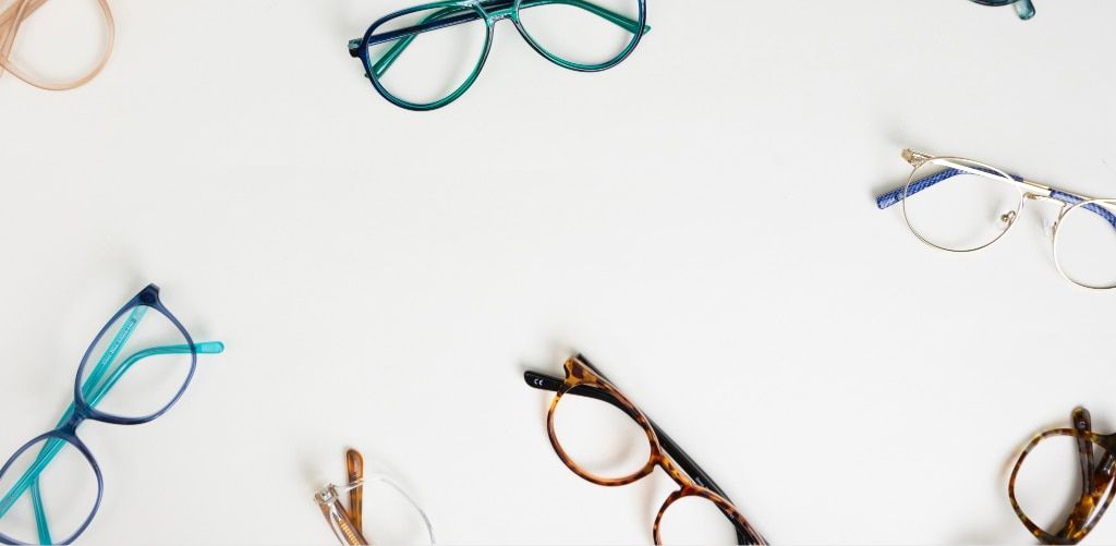 A selection of eyeglass frames in various shapes and colours, including blue, teal, tortoiseshell, gold and clear styles, arranged around the edges of a white background. The minimalist layout leaves ample space in the centre, creating a clean product dis