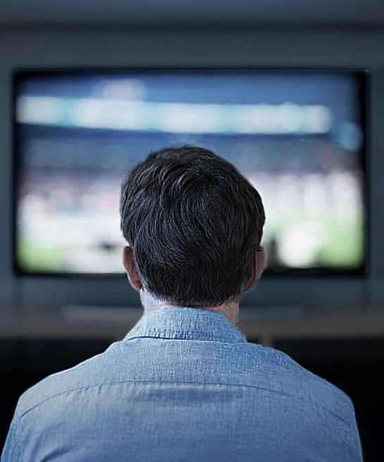 A close-up of the back of a man with short dark hair wearing a blue shirt, sitting close to a wall-mounted widescreen TV that appears blurred in the background.