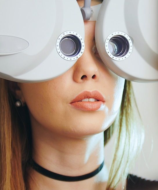 A young blonde woman looking through a phoropter during an eye exam, as though having her vision checked.