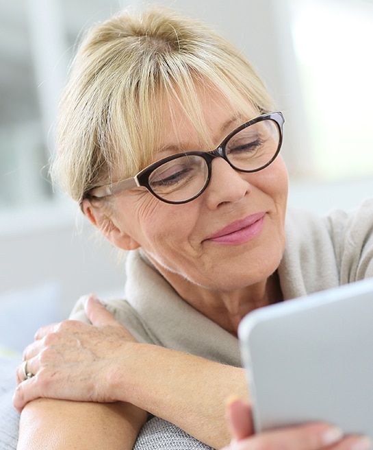 A close-up of an older woman with blonde hair, tied back, and a fringe. She is wearing black cat-eye glasses and a grey short-sleeve sweater, smiling as she holds a tablet in her right hand while her left hand rests on her right shoulder. She is in a brig