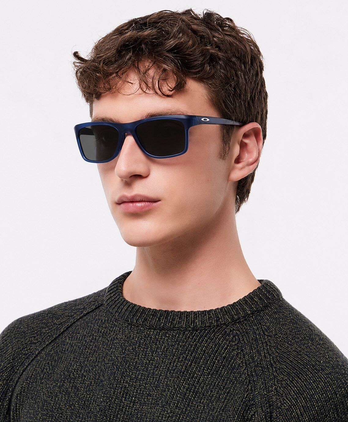A young man with short curly brown hair wears blue rectangular Oakley sunglasses and a dark sweater, looking slightly to the side with a neutral expression against a plain background.