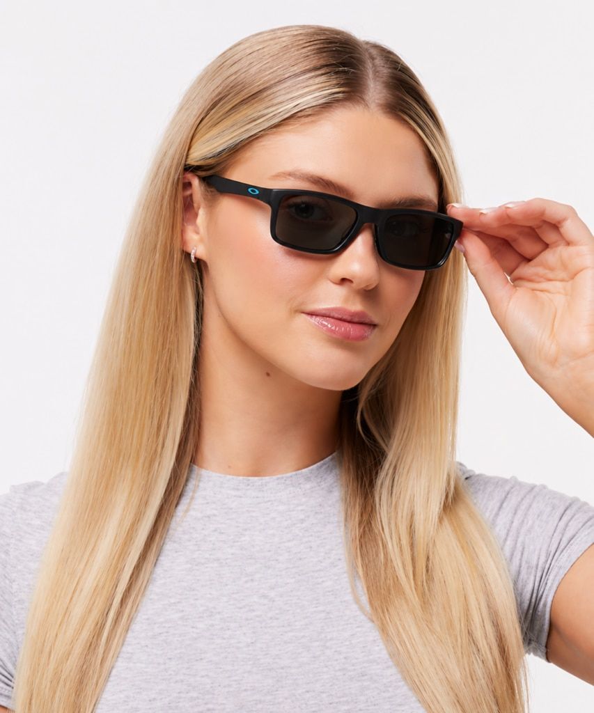 A blonde woman wearing Oakley Plank 2.0 rectangular black sunglasses and a light grey fitted t-shirt poses against a clean white studio background, gently adjusting the frame with one hand.