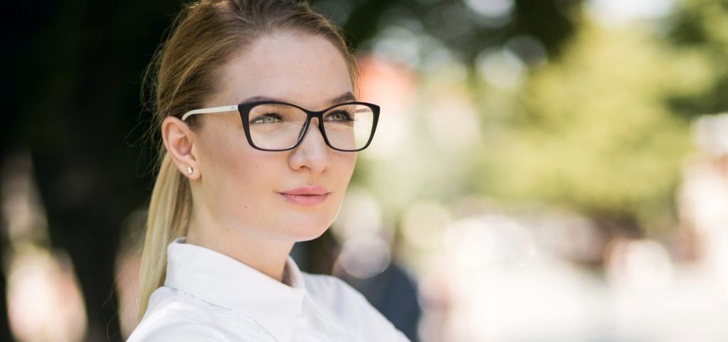 A young blonde lady wearing subtle cat-eye glasses which are black at the front and white along the temples. Her hair is in a ponytail and she is wearing a white shirt. She is looking into the distance and is in a bright outdoor setting which is blurred i